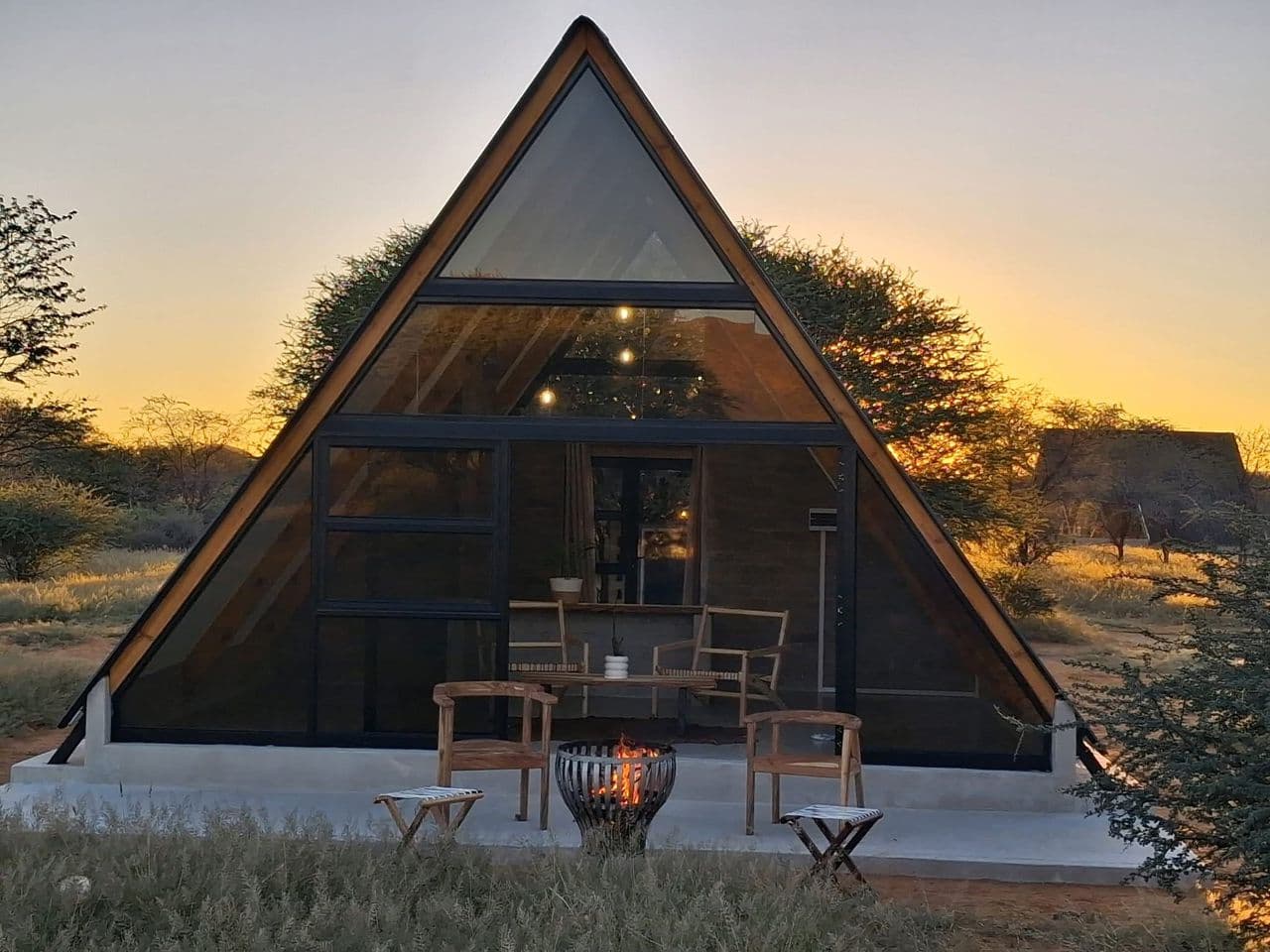 Modern A-frame glass cabin with a fire pit and outdoor seating at golden sunset.