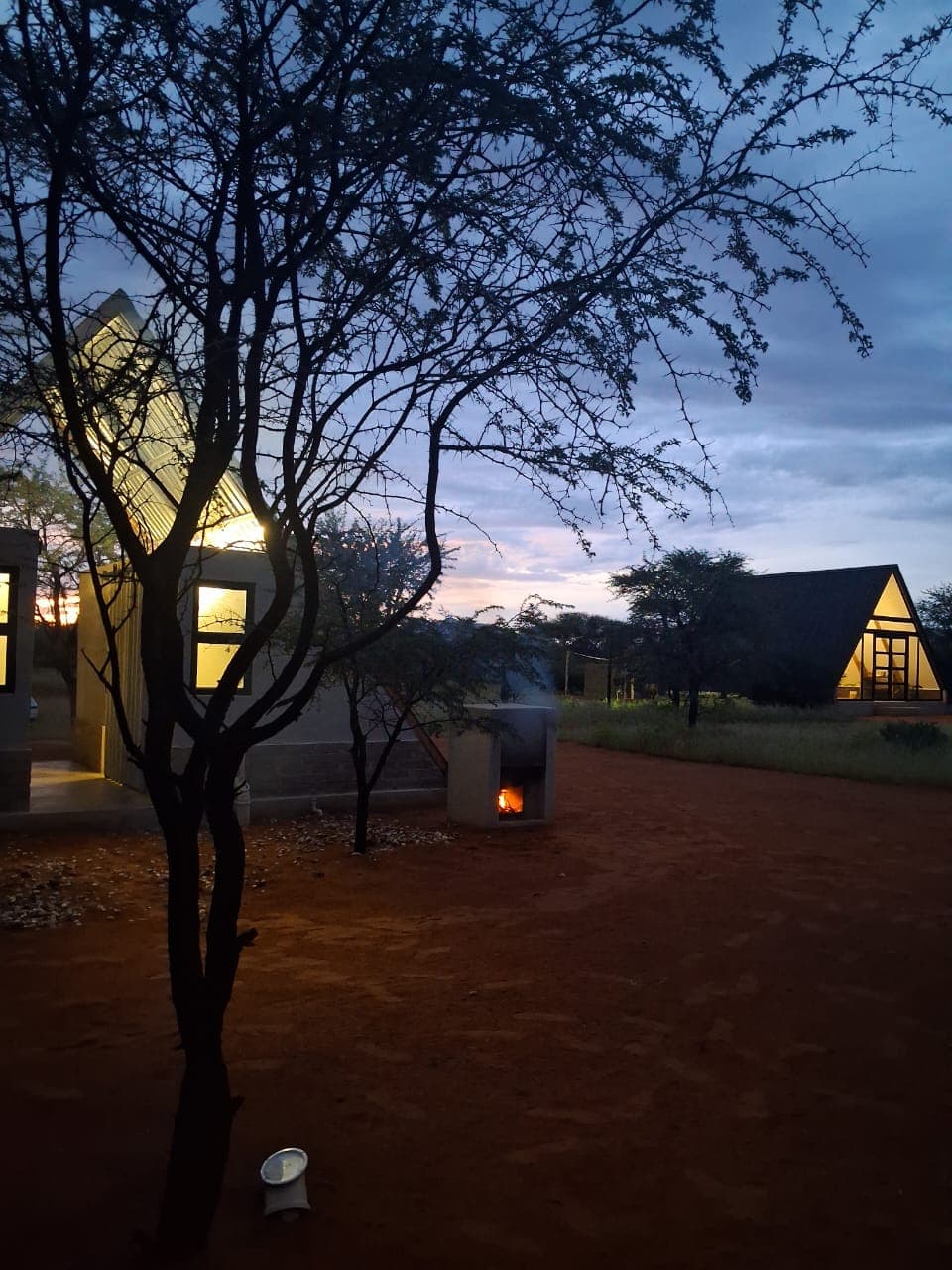 Illuminated A-frame cabins in a desert landscape at twilight with a small outdoor fire.