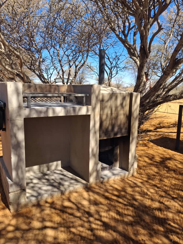 Outdoor concrete grill and wood-fired heater under sparse trees in a sunny, sandy landscape.