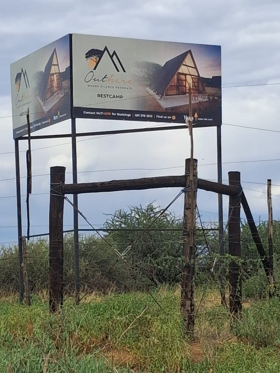 Billboard for Outhere Restcamp featuring an A-frame cabin, positioned behind a weathered wooden fence.
